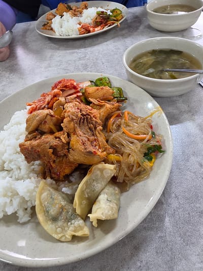 An overhead shot captures a close-up of a Korean meal laid out on a gray, textured surface, possibly a table. In the foreground, a large, off-white plate displays a generous portion of white rice topped with spicy braised chicken and kimchi. To the right of the rice is a pile of glass noodles with mixed vegetables, and at the bottom of the plate are several fried dumplings. A white bowl filled with a clear broth containing green leafy vegetables and some other ingredients is visible in the upper right quadrant of the image. Another similar bowl, partially obscured, is located behind the first. In the background, a smaller plate holds a portion of rice with kimchi and what appears to be some sort of stir-fried meat. The scene is likely within a casual dining establishment or a home setting in Daejeon, South Korea, given the typical presentation of Korean home-style meals.
