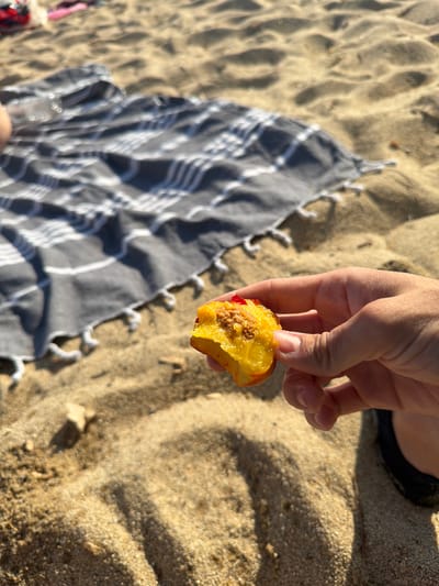 A hand holds a piece of a bitten peach over a sandy beach. In the background, a striped towel is laid out on the sand. The environment is a beach, likely in Sithonia Municipal Unit, Greece, given the sandy texture and the sunlight casting shadows.