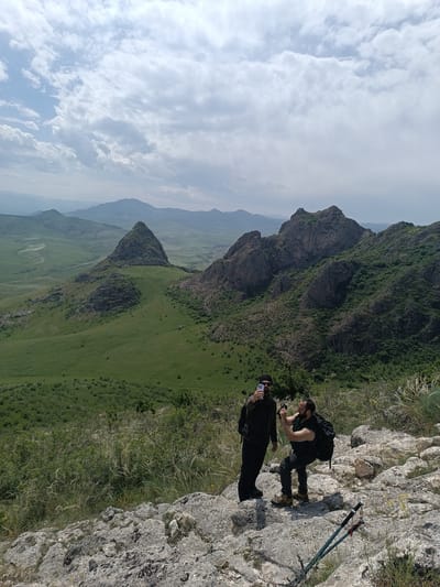 Two men are standing on a rocky outcrop in a mountainous landscape. One man, wearing a black jacket, black pants, and sunglasses, is holding up a smartphone, presumably taking a selfie. The other man, wearing a black t-shirt, grey jeans, and brown boots, is crouching and appears to be taking a picture with a camera. Both men are wearing backpacks. Hiking poles are visible on the ground near the edge of the outcrop. The background features rolling green hills, rocky mountains, and a sky with patchy clouds. Based on the context, it is likely that the location is in the vicinity of Hüseynbəyli, Azerbaijan.