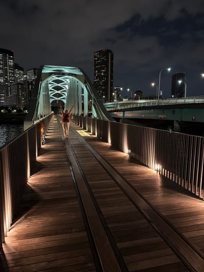 A person stands on a wooden bridge at night in Koto City, Japan, with their arms raised. The bridge, which appears to be part of a pedestrian walkway, is illuminated by warm lights embedded along the railings, casting long shadows on the wooden planks. The walkway is flanked by metal railings. In the background, a network of city lights from skyscrapers and another bridge are visible against a dark, cloudy sky. A river flows beside the bridge. The person is wearing light-colored pants and a reddish-brown top, and has a dark bag slung across their body.