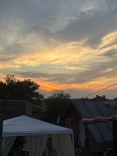 This image captures a sunset over a rural scene in Sredets, Bulgaria. The sky is filled with dramatic clouds, painted in hues of orange, yellow, and gray. In the foreground, a white gazebo with sheer curtains is partially visible on the left, suggesting an outdoor gathering or event. Behind it, several rustic buildings with corrugated metal roofs are scattered across the landscape. Trees with dark foliage are silhouetted against the vibrant sky. Electrical wires stretch across the scene, indicating a rural infrastructure. The overall impression is one of a peaceful evening in a Bulgarian village.