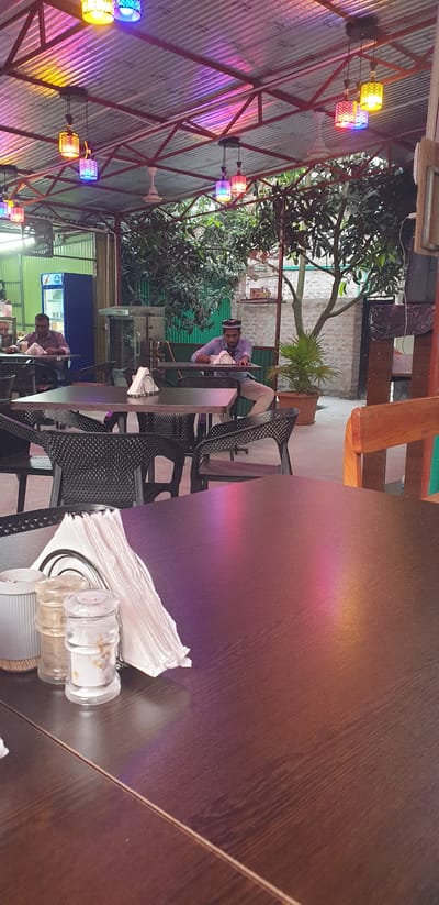 The image depicts an outdoor seating area of a restaurant, likely located in Chor Sahapur, Bangladesh, given the contextual information. The scene is illuminated by colorful pendant lights hanging from a corrugated metal roof, casting pink, purple, and yellow hues across the space. Several dark plastic chairs surround dark-colored tables.

In the background, two men are seated at tables. The man on the left, wearing a purple shirt, is looking down at his table. The man on the right, also in a purple shirt and wearing a cap, is leaning forward on his table, looking towards his left. A white napkin holder is visible on the table in the foreground, along with salt and pepper shakers. Beyond the seating area, there are trees and a brick wall. A blue refrigerator is partially visible in the left background. The overall impression is of a casual, open-air dining establishment.