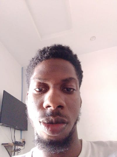 This media file is a close-up selfie of a Black man, taken indoors. The man has dark, curly hair and a goatee. His eyes are open, and he is looking towards the camera with a neutral expression. The background is a plain, white wall, with a portion of a television mounted on the wall visible on the left side of the frame. There is also a ceiling light fixture visible. The lighting suggests it is daytime, with the light source possibly coming from a window or overhead lights. The setting appears to be a home or an office. There is no visible text in the image. The overall impression is that of a casual, personal photograph.