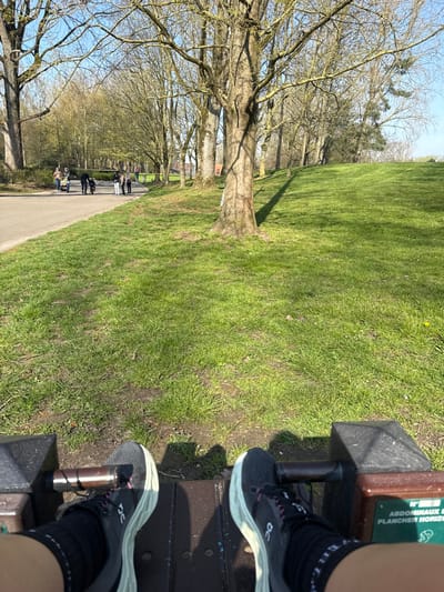 The image depicts a first-person view from an individual operating an outdoor exercise machine in a public park in Wattrelos, France, under clear, sunny conditions. The subject's feet, wearing black athletic shoes with light green/white soles and pink accents (brand 'On'), are positioned on the footrests of a dark-colored machine. A partially visible green plaque on the machine's right side displays French text, including "ABDOMINAUX" and "PLANCHER HORIZ".

The immediate foreground consists of green grass with some sparse patches of soil, illuminated by strong sunlight casting distinct shadows. A paved asphalt path curves through the midground on the left, alongside which a group of approximately three adults and two children, including one in a stroller, are congregated. Further left on the path, an adult and a child walk away from the viewer.

The background is dominated by numerous mature deciduous trees with bare branches, characteristic of late winter or early spring. These trees line the path and extend up a gentle grassy incline to the right. A clear blue sky is visible above the tree line, and a building with an orange-brown roof is faintly discernible through the distant trees on the left.