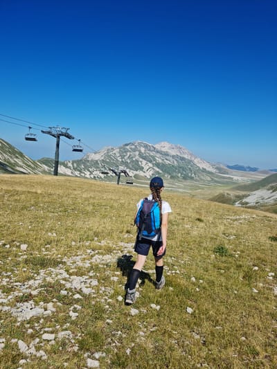 In a high-altitude, grassy mountain landscape under a clear blue sky, a young person with braided hair wearing a blue backpack, white t-shirt, dark shorts, dark socks, and hiking boots stands facing away from the camera. The terrain is covered in dry, yellow grass and scattered rocks. In the background, a ski lift with multiple chairs is visible, extending across the frame. Beyond the ski lift, majestic, rugged mountains with visible rock formations and patches of vegetation rise against the horizon. The scene suggests a summer day in a mountainous region, possibly the Gran Sasso mountain range near L'Aquila, Italy, known for its ski resorts and hiking trails.
