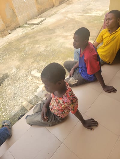 Three young people are sitting on a tiled porch outside a building with a textured, ochre-colored wall. The person in the foreground, on the left, is wearing a colorful floral-patterned t-shirt and dark pants. Their left arm is resting on the tiled floor, palm down. To their right, another young person is wearing a blue and red t-shirt and dark pants. Their right arm is also resting on the tiled floor. Further to the right, a third young person is wearing a bright yellow polo shirt. They are looking to the right. The ground outside the porch is rough and uneven, with some patches of greenery. A mop handle is visible behind the person in the yellow shirt. The scene appears to be outdoors, in what could be a residential or community area.