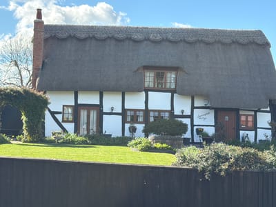 The image depicts a quaint, thatched-roof cottage in Malvern Hills, United Kingdom. The cottage, named "Summerfield Cottage," features white walls with black timber framing. A tall brick chimney rises from the left side of the roof. A small dormer window is visible in the thatched roof. The front yard is a well-maintained lawn with various plants and flower arrangements. A dark-colored wooden fence is in the foreground, partially obscuring the view of the garden. The sky is blue with scattered white clouds. The scene appears to be captured during daylight hours.
