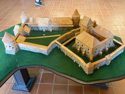 A detailed scale model of Trakai Island Castle, located in Trakai, Lithuania, is displayed on a dark-brown, curved table. The model features a sprawling complex of wooden and stone structures, including several towers with conical roofs, a large central courtyard, and extensive defensive walls. The model is situated on a green, grassy base, designed to represent the surrounding landscape. The table is placed on a floor tiled with reddish-brown squares. In the upper left portion of the image, the lower legs and feet of a person wearing athletic shoes are visible, indicating their presence near the display. The lighting appears to be natural, coming from above.