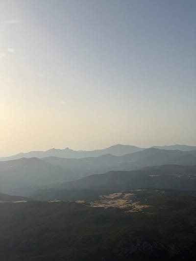 This is an aerial photograph of a mountainous landscape near Monte/Monti, Italy. The image features multiple layers of hazy, silhouetted mountains fading into the distance. The sky is a pale, clear blue, transitioning to a soft yellow near the horizon, suggesting either early morning or late afternoon light. The foreground is a dark, densely vegetated mountainside, with patches of lighter, dry vegetation or clearings visible on a plateau further into the scene. There are no people or man-made structures immediately discernible in the image, focusing solely on the natural topography.