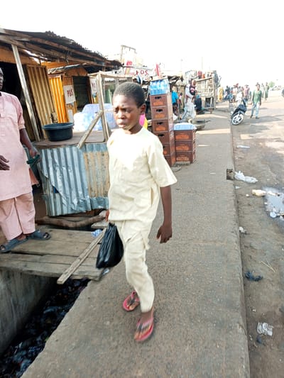 The image captures a street scene in Gadao, Nigeria. The foreground features a young boy walking towards the camera, dressed in a cream-colored traditional outfit and flip-flops. He is holding a black plastic bag in his right hand. To his left, an adult, also in traditional attire, stands partially in the frame, wearing a pale pink outfit and sandals.

Behind them, a row of small market stalls or shops are visible. These structures appear to be made of corrugated metal and wood, with some displaying wares like bottled water. Several crates of "MALTINA" beverages are stacked outside one of the stalls. A black basin sits on a raised wooden platform.

Further down the street, other people are seen walking and interacting, some carrying goods. A motorcycle is parked on the side of the road. The ground is paved, with a drainage ditch running alongside the walkway, filled with debris and trash. The overall impression is of a bustling marketplace on a clear, sunny day, likely in the late morning or early afternoon given the bright lighting and the shadows. The scene suggests everyday life and commerce in a Nigerian town.