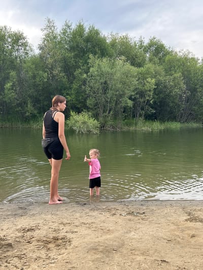 In this horizontal, eye-level shot, two females are at the edge of a body of water, with a sandy shore on the right and a dense treeline on the left. The woman in the foreground, wearing a black tank top and black shorts, stands with her left leg submerged in the water. Her posture suggests she might be engaged in a stretch or yoga pose. To her right, further into the water, a young girl in a pink t-shirt and black shorts stands with her hands outstretched towards the woman, seemingly interacting or playing. The water is a murky green, with ripples suggesting a gentle breeze. The treeline is lush and green, with the sky above appearing overcast. The scene evokes a sense of a casual outdoor activity, possibly a family outing or playtime on a warm, cloudy day. The location is identified as Angarsk, Russia. There is no visible text in the image.