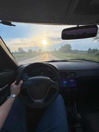 The image shows the interior of a car from the driver's perspective, with the steering wheel prominent in the foreground. The driver's left hand is on the steering wheel, and their right arm is visible, wearing a watch. Blue jeans are visible on the driver's lap. The dashboard features a car stereo with a "Bluetooth" display and playback controls. The car is in motion, as indicated by the road visible through the windshield and the sun low in the sky.
The setting is a road in a rural area, likely near Кондратово, Russia. Trees and fields are visible on either side of the road. The sun is setting, casting a warm glow and creating a slight lens flare effect. The sky is blue with wispy clouds. The overall mood is one of a peaceful drive during golden hour. The time of day is sunset.