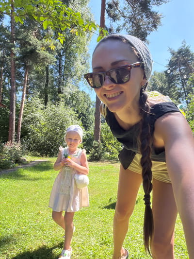 In a sunlit, grassy yard in Udelnaya, Russia, a woman with a bandana and sunglasses smiles into the camera, her long braid trailing down her back. She is wearing a black top and light-colored shorts. To her left, a young girl in a striped dress and a white headscarf holds a plastic bottle. The background is filled with lush green trees under a clear blue sky.