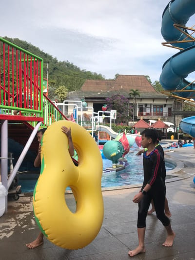 The image shows a water park located at the coordinates -7.882, 112.515 in Batu, Indonesia. A young boy is carrying a large yellow inflatable ring shaped like an "8". The ring is large enough to cover his body. He is walking towards the right, and there is another boy wearing black swimwear standing with his right hand outstretched to the right. There are other people and inflatable figures in the distance. The water park features colorful slides, play structures, and a blue pool with a cloudy sky overhead.  The scene suggests a fun, carefree atmosphere, with children enjoying the water park facilities.