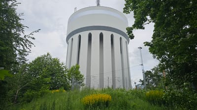 A tall, white cylindrical water tower with grey banding and arched vertical indentations stands prominently in the center of the frame, surrounded by lush green trees and foliage. The sky is overcast. In the foreground, tall grasses and yellow wildflowers are visible. A chain-link fence runs along the base of the tower, separating it from the grassy area. A cell tower or transmission tower is visible to the right of the water tower. This is the Roxnäs water tower in Linköping, Sweden.