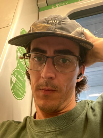 The image shows a close-up, selfie-style shot of a man wearing a camouflage baseball cap with "HUF" embroidered on the front, dark-rimmed glasses, and a green crew-neck sweatshirt. He has dark brown hair and is holding his right hand behind his head. A black wireless earbud is visible in his right ear. He is in what appears to be public transportation, possibly a train or metro car.

Visible on the wall behind him are two green circular stickers. The top sticker has a symbol of a plug with an arrow pointing down and text that reads "220V". Below this, the second sticker has a plug symbol with an arrow pointing down and the text "Endolla't" which means "Plug it in" in Catalan. Below this, there is further text in Catalan, including "Recorda" which means "Remember". The context of these stickers suggests the man is in a location within Sant Cugat del Vallès, Spain, where electrical outlets or charging points are available and a reminder to plug in devices is displayed. The background wall is light-colored with some grey paneling and a window. The lighting is warm and appears to be artificial.