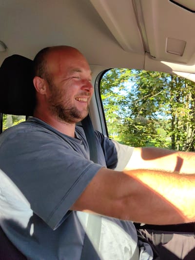 A man with a shaved head and a beard is driving a car, smiling with his eyes closed. He is wearing a grey t-shirt. Sunlight streams in through the car window, illuminating his arms and casting shadows on his shirt. Outside the window, green trees and a blue sky are visible, suggesting it is a sunny day. The interior of the car, including the headliner and a portion of the seat, is also visible. The image appears to be a candid shot, capturing a moment of joy or relaxation. The location is specified as Shchomyslitsky Sel'sky Sovet, Belarus.