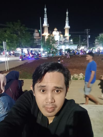An image captured at night in Kaliwungu, Indonesia, depicts a male subject in the foreground taking a selfie, with a brightly illuminated mosque and a bustling public area in the background.
The primary subject, an adult male, occupies the lower half of the frame. He has dark hair, dark eyes, and a light mustache and goatee. He is wearing a black collared shirt or jacket and is looking directly into the camera. His expression is neutral with slightly pursed lips. To his left, partially visible, is another individual wearing a light brown hijab and a dark red garment, with a blue denim garment also partially visible.
In the mid-ground and background, the scene is a public square or park area, illuminated by various artificial lights. A prominent white and green mosque with two tall, slender minarets topped with finials and multiple ornate domes is the central architectural feature. Its facade is brightly lit. In front of the mosque, a large number of people are congregated. The ground appears to be a mix of paved areas and packed earth. Several children are visible, some playing with small, illuminated ride-on vehicles (scooters or toy cars). A sign with partially visible text "KACAMA" (likely "Kacamata" meaning eyeglasses) is seen to the left. Trees with green foliage are present, illuminated by the ambient light. Power lines and utility poles are visible against the dark night sky. To the right, a male figure wearing a blue t-shirt and light shorts is walking, his face blurred by motion. The overall scene suggests a lively evening gathering.