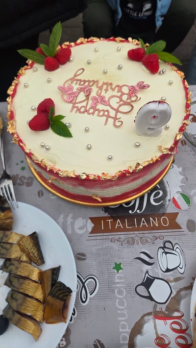 A birthday cake with "Happy Birthday" written on it in rose gold lettering and a number "9" candle is placed on a table with other food items and a checkered tablecloth. The cake is decorated with raspberries, mint leaves, and gold leaf accents. To the left of the cake, a plate of sliced smoked fish is visible, along with a fork. The background shows a person wearing a black t-shirt with "Little League" printed on it. The scene is set outdoors, suggesting a celebration.