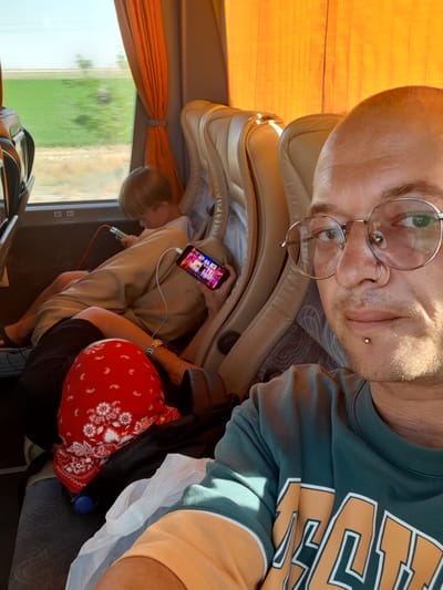 The image shows a selfie taken from the perspective of a person with a shaved head, wearing glasses and a nose ring, on a bus. The person is looking directly at the camera. In the background, another person, a child with blonde hair, is seated and appears to be looking at a mobile phone. A second mobile phone, playing a video, is held by an unseen hand. The bus interior is visible, with beige seats and orange curtains. Outside the window, a blurred green field and a blue sky are visible. The setting appears to be a travel scenario on a bus, potentially within or near Karatay, Turkey, based on the provided location information. The image captures a moment of transit, with individuals engaged with their electronic devices.