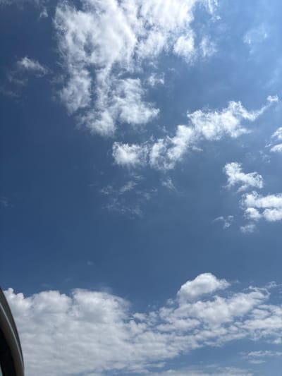 This is a vertical, eye-level, full-shot photograph taken from a moving vehicle looking upwards at a clear blue sky with scattered white clouds. A curved, dark gray object, which appears to be the frame of a car window or sunroof, is visible in the lower left corner of the image. The clouds are varied in size and density, with some appearing wispy and others more voluminous and puffy. The sunlight is bright, casting a soft illumination on the clouds, and the overall impression is of a pleasant, sunny day. The location is the city of Opatów, Poland.