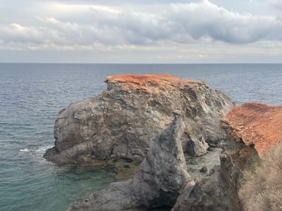 The image displays a rugged coastal scene in Cabo de Palos, Spain. In the foreground on the right, a reddish-brown cliff edge with sparse, dry, light brown vegetation slopes downwards. The midground is dominated by a large, dark grey rock formation emerging from the sea on the left, its flat top covered by a distinct layer of reddish-brown earth or rock. To the right of this large formation, a jagged, spire-like dark grey rock pinnacle rises sharply from the water. The sea around these rocks is a clear, greenish-blue, indicating shallowness or light penetration, with small waves breaking at the base of the rock structures. Further out, the open sea extends to the horizon, appearing a deep, dark blue. The sky above is overcast, filled with layered, grey and white clouds, suggesting a cloudy day. No people or man-made objects are visible.