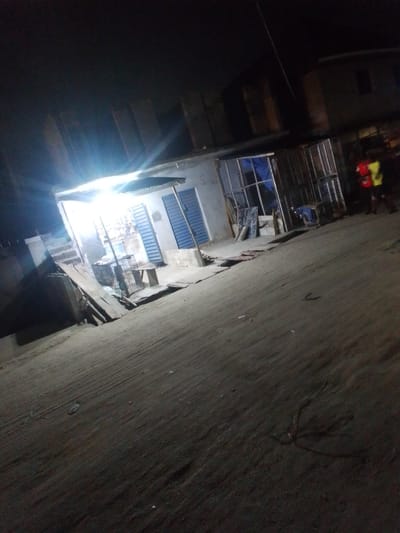 This nighttime photograph captures a brightly lit street-side stall or shop in what appears to be an urban or semi-urban area, likely Awoyaya, Nigeria. The image is taken at a tilted angle, enhancing the dynamic feel of the scene.
The central subject is a small, single-story building with a corrugated metal roof, intensely illuminated by bright artificial lights, possibly fluorescent, which cast a strong glare. Inside and directly in front of the structure, various items are visible on shelves or display racks, although their specific nature is obscured by the brightness. Wooden planks lean against the facade, and a small wooden table stands nearby. On the right side of the stall, there's an open structure featuring metal grates or cages and some blue coverings or tarps.
To the far right, a person is partially visible, wearing a brightly colored yellow or orange vest or shirt, suggesting a worker or a pedestrian. Their activity is unclear. The ground in the foreground and surrounding the stall appears to be unpaved, possibly dirt, sand, or a rough concrete surface, with some scattered debris. In the background, darker, less defined structures of other buildings can be vaguely seen under the dark night sky.
The time of day is clearly night, with the artificial lights providing the sole illumination for the immediate area. There are no notable weather conditions discernible, and no visible text or signage is legible in the image. The scene suggests a functional, bustling area, possibly a local market or commercial spot, operating after dark.