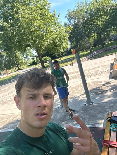 The image is a selfie taken outdoors, possibly in a park setting in Elhovo, Bulgaria, based on the text on the t-shirt of the other individual in the photo. The primary subject is a young man wearing a green t-shirt. Another young man is further away in the background, wearing a green and white t-shirt and blue shorts. He is standing near a metal exercise station. In the background, there are trees and a concrete surface that appears to be a walkway or a park area. Part of a wooden bench is visible in the upper right corner, and some exercise equipment is nearby. The selfie taker has a finger pointing towards the other individual.