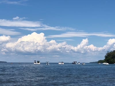 This image captures a serene view of Kempenfelt Bay in Barrie, Canada, under a vast, partly cloudy blue sky. The water is calm and deep blue, with gentle ripples suggesting a light breeze. Several boats are scattered across the bay, varying in size and type, including speedboats and pontoons. Some boats appear to be anchored, while others are in motion. In the distance, the shoreline is visible on the left, and on the right, a densely wooded area forms the backdrop, hinting at a natural park or conservation area. The overall atmosphere is one of peaceful recreation on a summer day.