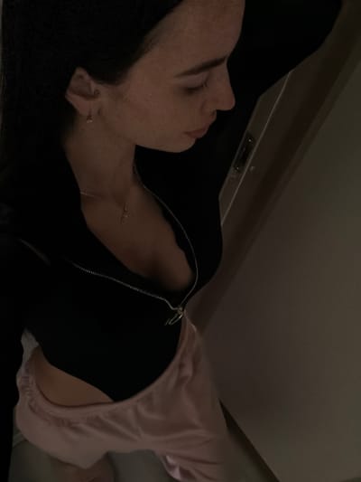 A dimly lit, high-angle interior photograph displays a female individual, located in Krasnaya Zarya, Russia. The subject is depicted from the mid-torso upwards, positioned in profile, with her dark hair falling behind her head. Her gaze is directed downwards and towards the right side of the frame. She is wearing a long-sleeved black top with a silver zipper, partially unfastened to expose the upper chest. Beneath the top, the waistband of light pink, loose-fitting pants, possibly satin, is visible. A delicate silver-colored necklace with a small, indistinct pendant is around her neck, and a small earring is present in her visible ear. Her right arm is raised, with the hand out of frame, suggesting she is leaning against an unseen vertical surface. The background consists of plain, light-colored walls, with a portion of a doorframe or architectural panel, possibly including a metallic latch mechanism, visible to the right of her head.