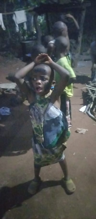 This image depicts a low-light, likely evening or nighttime, scene in Amokwue-Affa, Nigeria, featuring several children in a rustic outdoor setting.

The main subject is a young child in the foreground, facing the camera with a dramatic, wide-eyed expression and open mouth, hands raised above their head. This child wears a patterned dress or tunic, a mix of green, black, and white, and light-colored slip-on shoes. Dark markings, possibly paint or dirt, are visible on their face.

Behind the central figure, other children are present, including one in a light green shirt looking away from the camera. The ground is bare earth or dirt. In the dimly lit background to the left, white fabric, possibly laundry, hangs on a line or branch, near a large dark pot or container and some bottles. To the right, a pile of sticks or cut wood is visible. The overall scene suggests a candid moment of play or activity in a village environment. No text is visible in the image.