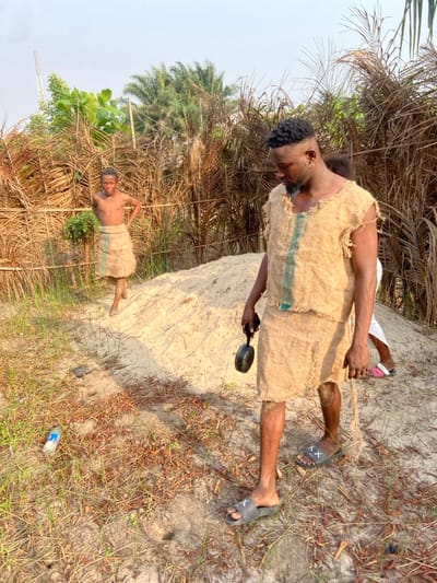 The image, taken in Igbologun, Nigeria, features two males outdoors on a bright, sunny day. The prominent figure in the foreground is a Black man, standing in a sandy area. He wears a rustic, light brown tunic made from what appears to be burlap or sackcloth, featuring a horizontal green stripe across the chest. He has a short, dark beard, an earring, and dark braided hair, and is looking downwards or to the side. On his feet are dark sandals with a distinct white "XX" symbol on the straps. He holds a black magnifying glass in his right hand.

Further back and to the left, a second, younger Black male stands shirtless, wearing a similar burlap-style garment as a skirt or loincloth, also with a green stripe. He is barefoot and looks towards the camera with a slight smile.

The scene is set on sandy ground with scattered dry grass and sparse vegetation. In the background, there is dense, tall tropical foliage, including what appears to be palm trees, indicating a rural or semi-wild environment. A discarded plastic bottle lies on the ground near the main subject's feet. The bright, clear light suggests it is midday or early afternoon. The burlap attire and magnifying glass hint at a themed event or performance, possibly portraying a survivalist or explorer character.