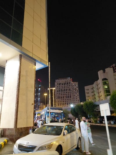 The image depicts an urban nighttime scene in Makkah Al Mukarramah, Saudi Arabia.
In the foreground, a white modern sedan is parked or stationary, angled slightly towards the right, occupying the lower center. To its immediate left, a brightly lit pillar with a marble-like finish supports a multi-story building featuring a beige tiled facade and dark windows. A black and yellow striped curb is visible at the bottom left.
In the midground, a diverse group of pedestrians, many dressed in white traditional garments (likely pilgrims), are visible near a blue bus with Arabic script, which appears to be a shuttle or public transport vehicle. A white parking barrier pole with red and white stripes is positioned behind the white sedan. To the right of the sedan, two men are standing; one with a beard, dressed in a white thobe, is holding a white bottle, looking towards the left. The other man, also in white attire, is partially obscured. Further to the right, a white utility pole with a rectangular sign attached is present.
The background features a dark night sky above a cityscape of illuminated, multi-story buildings. A prominent, light-colored building with numerous windows arranged in a grid pattern stands centrally. To its right, another building with green-tinted windows and an irregular facade is visible. Trees and foliage are interspersed at street level between the foreground and background structures. Streetlights illuminate various parts of the scene.