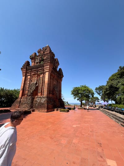 A man wearing a white robe and glasses stands in the foreground, facing left, on a terracotta-tiled plaza. In the middle ground, a tall, weathered, multi-tiered brick tower stands prominently. Beyond the tower, several people walk across the plaza, and lush green trees dot the landscape. In the background, the bright blue sky meets the horizon, with a hint of the sea visible. This scene is of the Thap Nhan (Cham Towers) in Ward 3, Tuy Hoa City, Phu Yen Province, Vietnam.