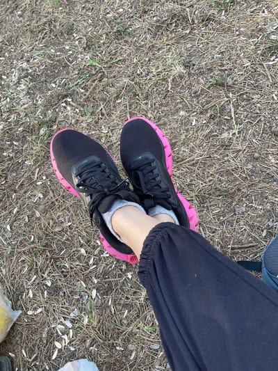 The image shows a close-up, overhead view of a person's legs and feet, wearing black athletic shoes with bright pink soles. The shoes are laced and appear to be on a person sitting outdoors, as indicated by the ground covered in dry leaves, twigs, and what appear to be sunflower seed shells. The person is wearing black sweatpants. The scene is outdoors, likely in a park or natural area, given the ground cover. The specific location in Arnavutköy, Turkey is not discernible from the image itself, as there are no identifying landmarks or contextual clues.