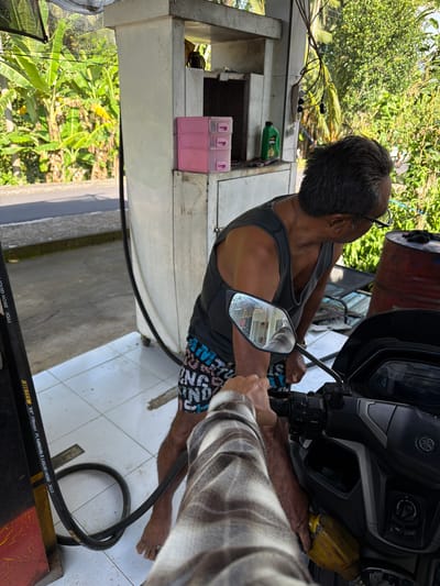In Selemadeg Timur, Indonesia, a person is refueling a scooter at a small, open-air gas station. The gas pump, a traditional column with a hose, is visible to the left. A middle-aged man, wearing a gray tank top and patterned shorts, stands by the pump, looking towards the scooter. The scooter's handlebars and dashboard are in the foreground, with a yellow fuel nozzle inserted into the scooter's fuel tank. The ground is tiled, and lush green vegetation, including banana trees, is visible in the background, suggesting a tropical environment. The scene is lit by natural daylight.