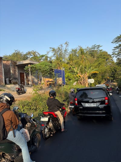 The image depicts a street scene in Madyocondro, Indonesia, characterized by a moderate traffic jam. Several motorcycles and a black car are visible, some with riders, suggesting they are either stationary or moving slowly. The environment is lush with vegetation, including trees and grass, indicating a rural or semi-rural setting. The time of day appears to be late afternoon, as suggested by the warm lighting and long shadows. The presence of a building to the left, a road, and overhead power lines further contribute to the urban or semi-urban context. There is a blue banner in the background with text, partially obscured, and a sign with text "ASRI" which may be a place name or advertisement. The sky is clear and blue.