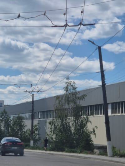 The image captures a street scene with overhead wires dominating the foreground against a backdrop of a cloudy blue sky. A dark, modern-looking car is parked on the right side of the road, its rear visible. Further down the road, a person wearing dark clothing is walking. Behind them, a building with horizontal window patterns is visible, along with trees and greenery. The scene appears to be outdoors during the daytime, with the lighting suggesting it might be late afternoon or early evening. The image is oriented vertically, with the sky and wires taking up most of the frame. The overall impression is a typical urban or suburban environment.