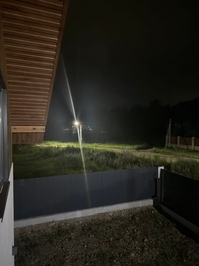 An outdoor, eye-level, nighttime shot shows a dimly lit rural scene from a porch. In the foreground, a dark grey metal fence runs horizontally across the image. To the right of the fence is a black metal gate. Beyond the fence is a patch of gravelly ground. A dark wooden ceiling with horizontal planks covers the top left portion of the image, part of a building's overhang. A single lamppost with a bright light is visible in the middle ground, illuminating a grassy area with some weeds. In the background, a dense line of trees forms a dark silhouette against the night sky. To the right of the trees, a brown brick fence with vertical supports is visible. The scene is devoid of people or animals. The overall impression is one of a quiet, residential outskirts of Malinovka, Russia, at night.