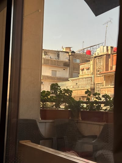 The image provides an exterior view captured from inside a structure in As-Salihiya Municipality, Syria, looking out over an urban residential environment under a clear sky. The immediate foreground reveals reflections on a window pane, showing parts of two dark plastic chairs. Below the visible window frame, a balcony railing holds multiple brown rectangular planters containing various green plants, including geranium-like foliage. Beyond the balcony, a dense arrangement of multi-story buildings is visible. These structures feature concrete or masonry construction, with details such as balconies, some enclosed with glass, and windows with security bars or shutters. Rooftops display typical urban infrastructure, including multiple satellite dishes, at least two prominent red cylindrical water tanks, air conditioning units, and a tall, multi-element television antenna on the right. Clothes are observed hanging on a clothesline on an upper balcony of a building on the left. The scene is illuminated by direct sunlight, creating distinct highlights and shadows across the building facades.