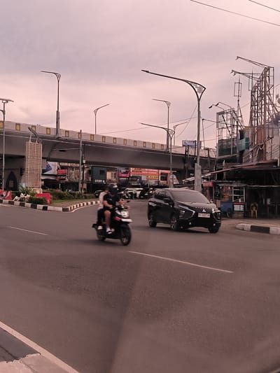 The image shows a street scene in Banda Aceh, Indonesia, likely during the late afternoon or early evening, given the lighting conditions. A black Mitsubishi Xpander is driving on the right side of the road, and a motorcycle carrying two people is on the left. In the background, there's an overpass, buildings, and streetlights. The sky has a pinkish hue. The general appearance of the buildings and street infrastructure suggests an urban setting.