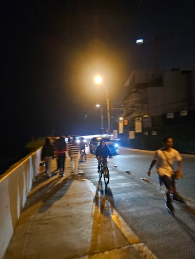 The image depicts a nighttime scene in Miraflores, Peru. Several people are present, some standing near a wall, and one running on the street near a cyclist. A bright street light illuminates the area. The presence of parked cars and buildings with signage indicates an urban setting.