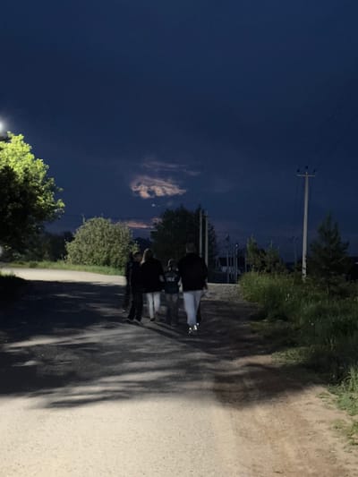 The image depicts an unlit road in Горный, Russia at night. A group of four people walk along the road's right side, casting long shadows due to a light source behind the camera. The road is bordered by vegetation on the right and trees on the left. Utility poles are visible in the background. The sky is dark with scattered clouds.