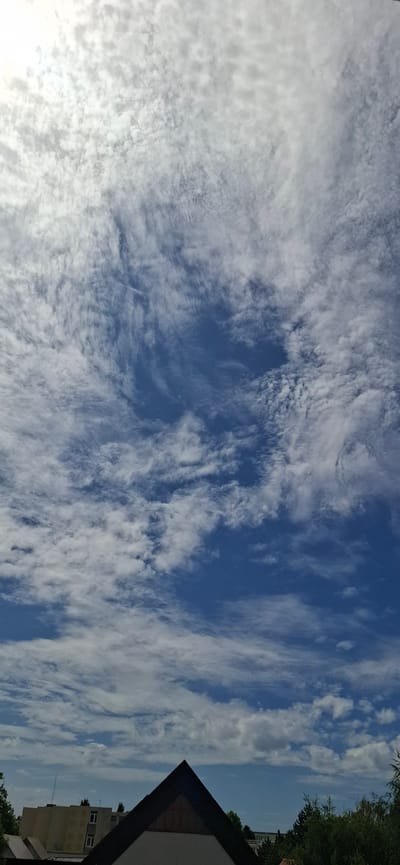 The image captures a sky filled with varied cloud formations, ranging from wispy cirrus clouds towards the top to more textured cumulus clouds lower down. The sky is a vibrant blue. In the foreground, the top of a dark, triangular-shaped roof is visible, with a red cross shape in the center. To the left of this roof, a multi-story building with a light-colored facade and windows can be seen. Further to the left and in the background, more buildings and trees are present. The scene appears to be an outdoor view from an elevated position, looking up at the sky and down onto a section of a city, likely Le Havre, France.