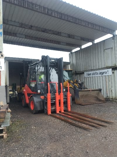The scene is an outdoor industrial or storage yard in Bitburg, Germany, under an overcast sky. The ground consists of dark gravel and dirt.

Dominating the midground is a large, heavy-duty forklift, primarily red and black, facing the viewer's left. It features four orange-painted forks extended forward. The brand "RINGO" is partially visible on its side, and it has red wheel hubs. Behind and to the right of the forklift, a yellow Volvo wheel loader is parked with its large, dirt-filled bucket resting on the ground.

The area is partially covered by a lean-to style roof constructed from corrugated metal panels and supported by visible metal I-beams, indicating an open-sided shelter. To the right, a wall composed of stacked, corrugated shipping containers or similar modular industrial panels forms a boundary. A white rectangular sign with black lettering is affixed to this container wall, displaying the German text: "Hier nichts lagern!" (meaning "Do not store anything here!"). To the far left, a portion of a concrete or light-colored building is visible with a dark, open entryway. No individuals are present, and no active operations are depicted. Several wooden pallets are stacked on the ground to the far left.