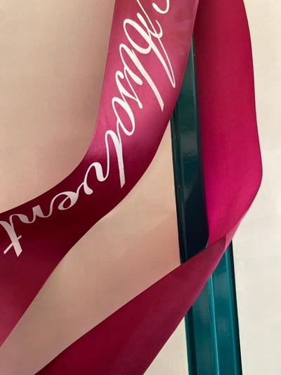 The image shows a close-up of a magenta ribbon with white text that appears to say "Březová". The ribbon is draped over a dark teal metal bar. The background is a textured, light-colored surface. The lighting suggests an indoor setting. There is no information about Bílina, Czechia in the image.