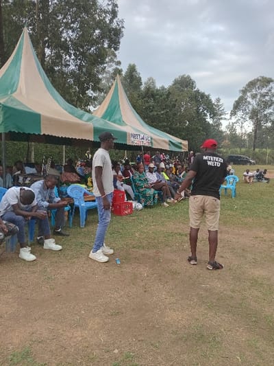 The image depicts an outdoor gathering in Chevaywa ward, Kenya. The scene features a group of individuals assembled on a grassy field under an overcast sky, with trees visible in the background.
Two large, green and beige striped tents are erected, providing shelter for the main crowd. A banner, partially visible on one tent, displays text including "WETU".
In the foreground, two adult males stand on a dirt patch surrounded by grass. The male on the left wears a light-colored t-shirt, dark baseball cap, blue jeans, and white sneakers, standing with his left side partially facing the viewer. The male on the right, with his back to the viewer, wears a black t-shirt with white text "MTETEZ WETU", a red baseball cap, khaki shorts, and dark sandals. A small blue object lies on the ground between these two individuals.
Beneath the tents, a large group of people, primarily adults, are seated on blue plastic chairs, facing towards the front of the gathering. Some red crates are visible near the seated attendees. To the far right, in the background, a dark vehicle is parked, and a smaller group of individuals are gathered around it on the grass. The overall scene suggests an organized outdoor community event or meeting.