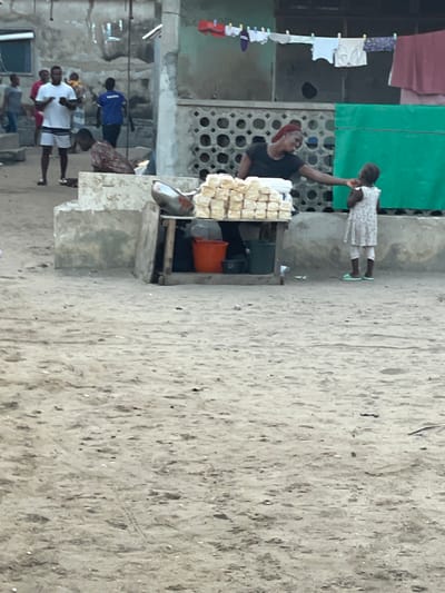 A woman with a red head covering stands behind a table filled with stacks of square, white objects, possibly bread or pastries. She reaches up to a young girl in a white dress. Clothes hang on a line in the background. Other people can be seen walking nearby. The scene appears to be set in a crowded outdoor location with a sandy ground. A green sheet is above the woman and child.