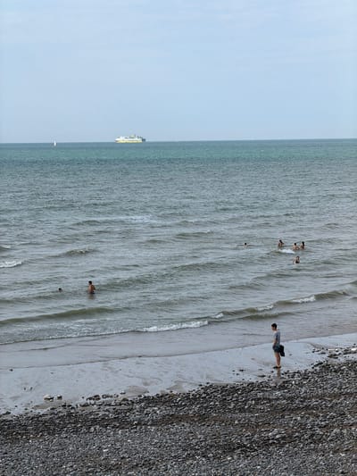The image depicts a view of the English Channel from the beach in Hautot-sur-Mer, France. In the distance, a large ferry is visible on the water, along with a small sailboat. Closer to the shore, several people are swimming and wading in the sea. A lone man stands on the wet sand near the edge of the water, facing the sea. The foreground is dominated by a pebble beach. The sky is overcast and a light breeze creates small waves on the water's surface.