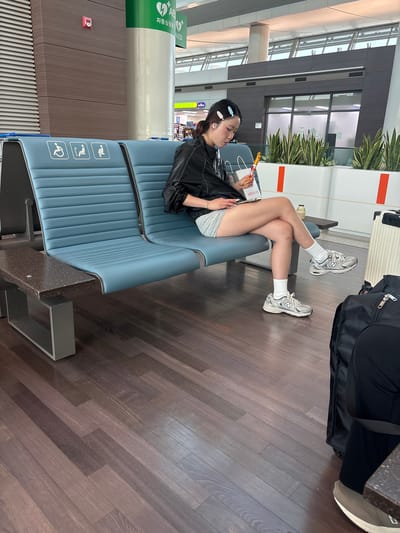 Incheon International Airport, South Korea. A young woman with light-colored hair clips is sitting on a blue airport waiting chair, with her legs crossed. She is wearing a black jacket over a white top, grey shorts, white socks, and silver athletic shoes. She is holding a pen and a small white notebook or device in her hands. Two other airport chairs are visible to her left, each with an accessibility icon. A dark brown granite table with a metal base is attached to the leftmost chair. In the background, a dark wooden floor leads to large windows and indoor plants. A black suitcase and a white wheeled suitcase are visible next to her.